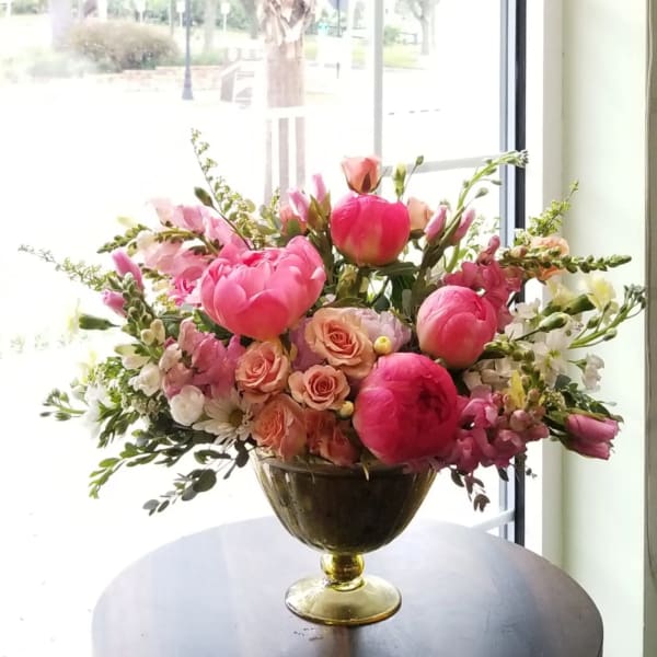 Low gold compote of bright pink peonies, peach roses, and white blooms on a table by a window