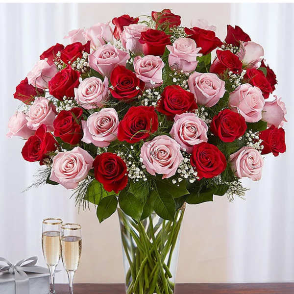 Tall clear vase of red and pink roses with baby's breath, next to two champagne glasses and a silver gift box.
