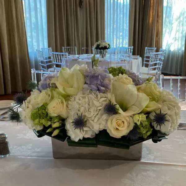 Low floral centerpiece with white roses, hydrangeas, and pale purple blooms in a rectangular vase