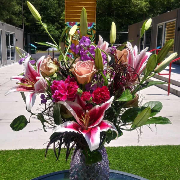Tall arrangement of pink lilies, peach roses, and bright carnations in a purple textured glass vase