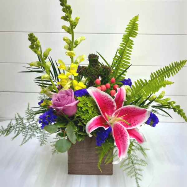 Mixed arrangement with pink lily, purple rose, yellow snapdragons, and ferns in a wooden cube.