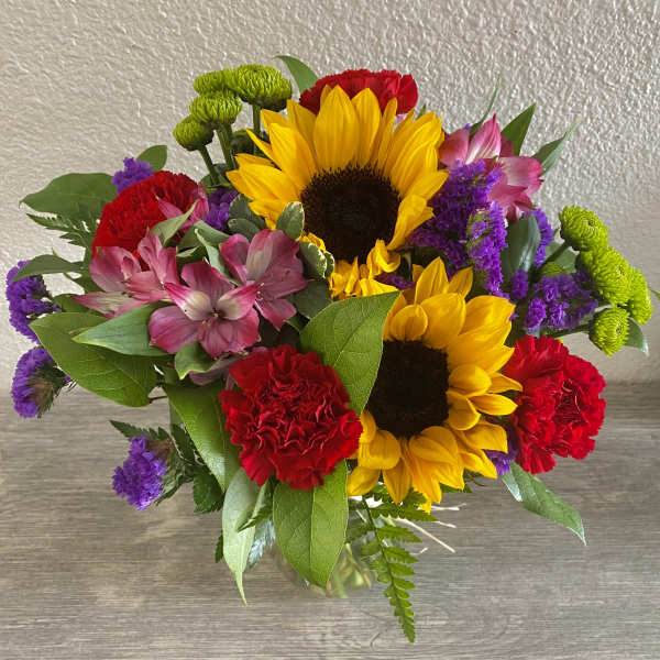 Bouquet with sunflowers, red carnations, pink alstroemeria, and purple filler in a glass vase