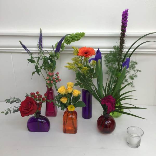 Small floral arrangements in colorful glass vases with roses, irises, and mixed blooms