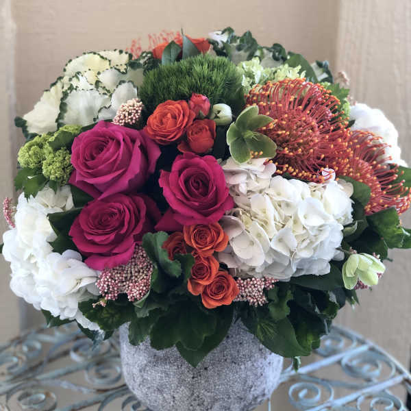 Low round arrangement of pink roses, orange spray roses, protea, and white hydrangeas in a gray pot
