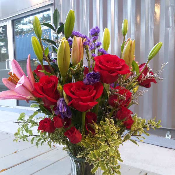 Tall bouquet of red roses, pink lilies, and purple accent flowers in a clear glass vase.
