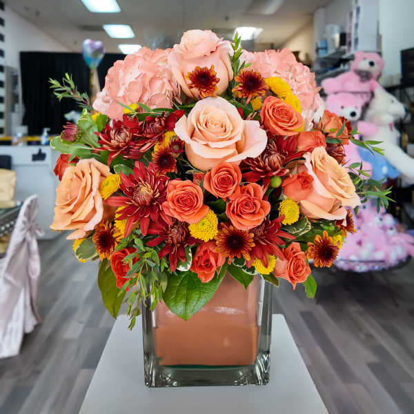 Peach and coral roses with red and yellow flowers in a square glass vase