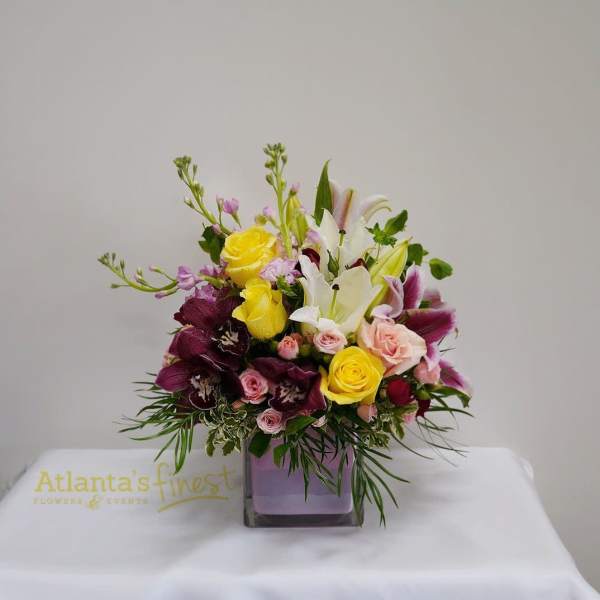 Mixed bouquet of yellow roses, white lilies, and pink blooms in a square vase