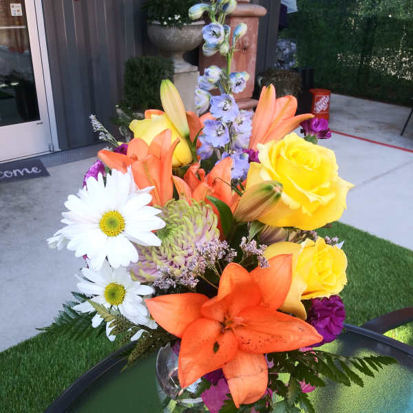 Bright mixed bouquet with lilies, roses, daisies, and purple accents in a glass vase