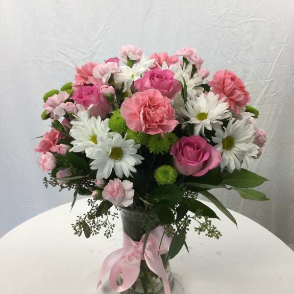Pink roses, carnations, and white daisies in a glass vase with a pink ribbon.