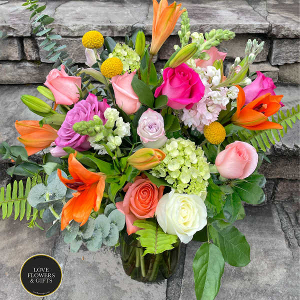 Colorful arrangement of pink roses, orange lilies, and mixed blooms in a clear glass vase.