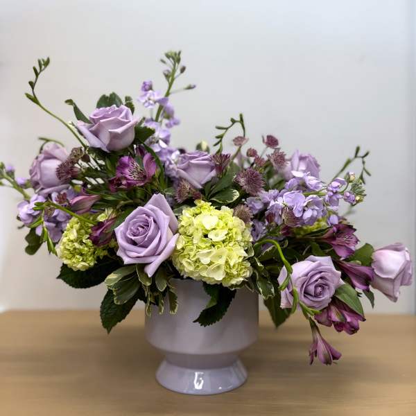 Lavender rose arrangement in a white pedestal vase with purple blooms