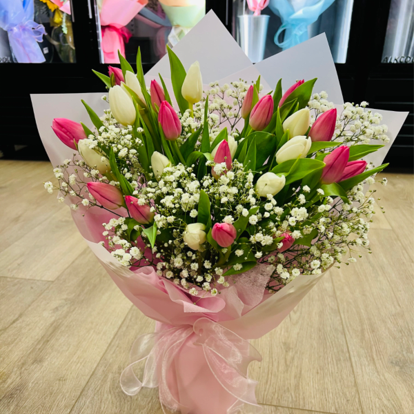 Hand-tied bouquet of pink and white tulips with small white filler flowers wrapped in pink paper and ribbon.