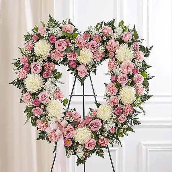 Heart-shaped standing wreath of pink roses, carnations, and white mums on a metal easel with a pink ribbon bow