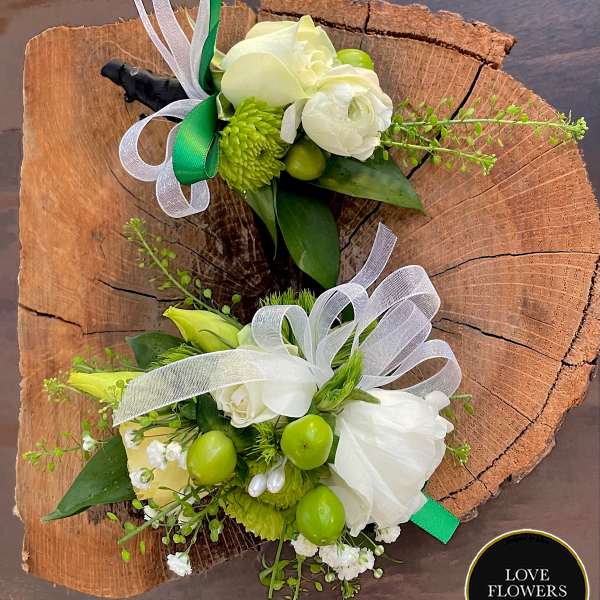 Pair of white and green floral boutonnieres with ribbons displayed on a rustic wood slice
