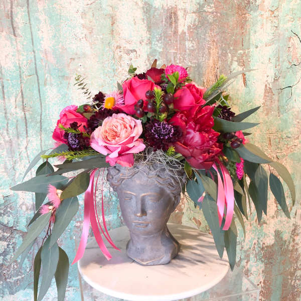 Pink and red floral arrangement in a gray bust vase with long ribbon streamers