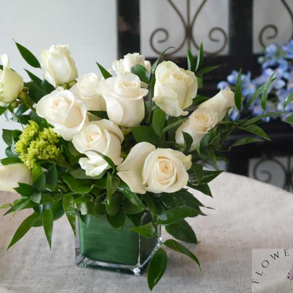 White roses arranged in a square glass vase with green foliage