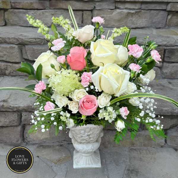Tall arrangement of white and pink roses with pink carnations and green hydrangea in a white urn vase.
