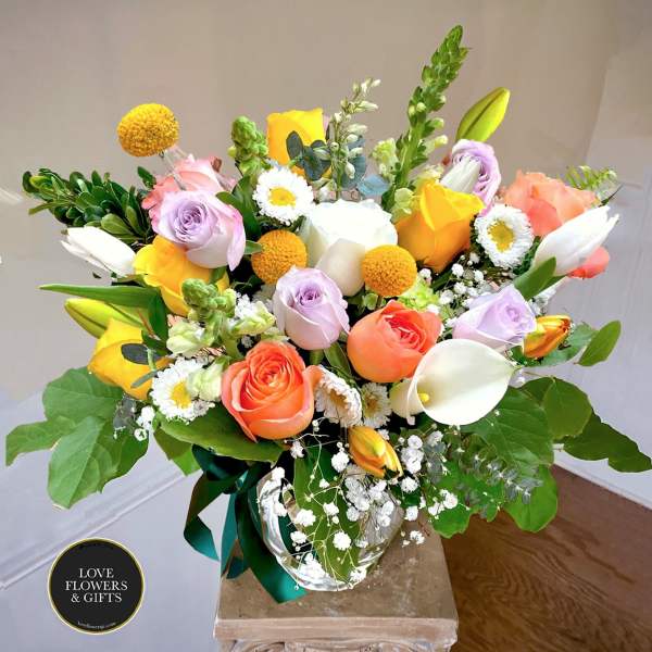 Mixed bouquet of yellow, peach, and lavender flowers with white calla lilies in a clear glass vase