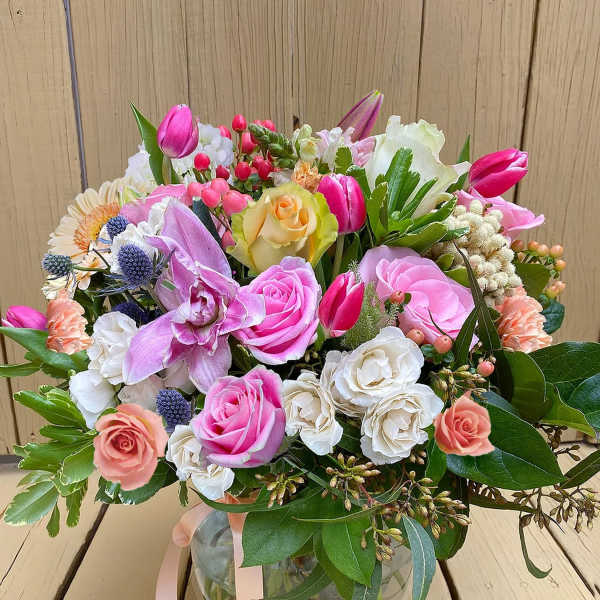Round glass vase of mixed pink, peach, and white flowers with ribbons on a wooden surface