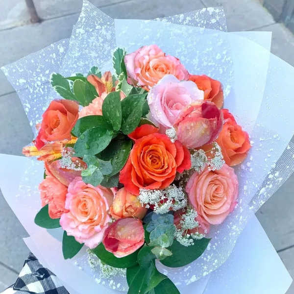 Handheld bouquet of orange and pink roses wrapped in white translucent paper.