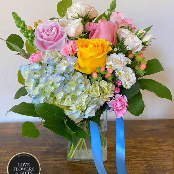 Mixed bouquet of pink and yellow roses, hydrangea, and carnations in a clear glass vase with blue ribbon