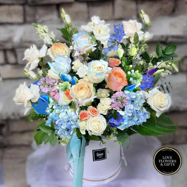 Pastel roses, hydrangeas, and mixed blooms arranged in a white hat box with aqua ribbon.