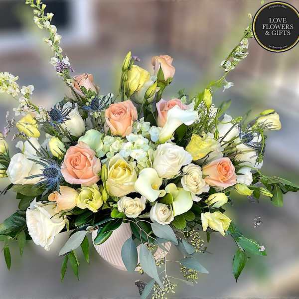 Low ceramic bowl of white, peach, and yellow roses, calla lilies, and mixed blooms in a lush arrangement.