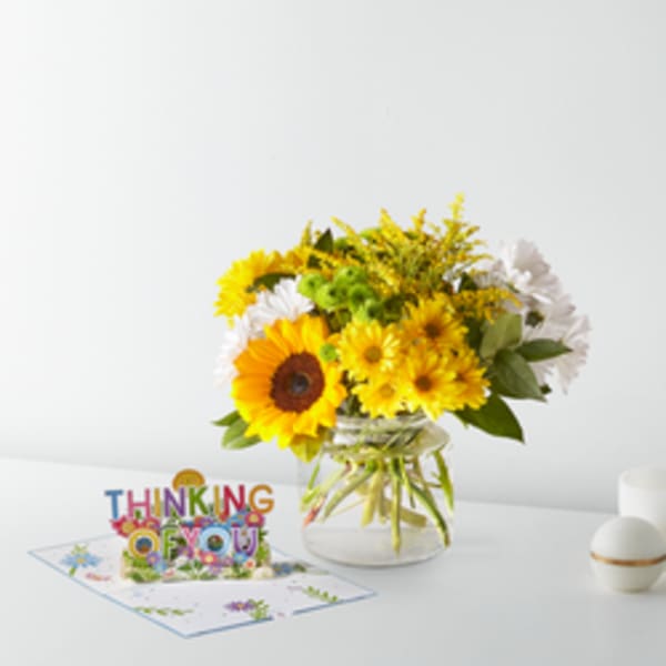 Yellow and white flowers in a glass vase beside a "Thinking of You" card