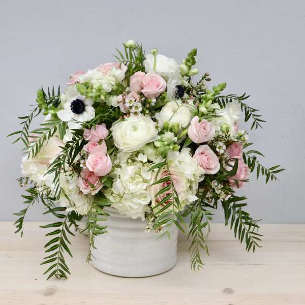 White and blush flower arrangement with trailing greenery in a white ceramic vase.