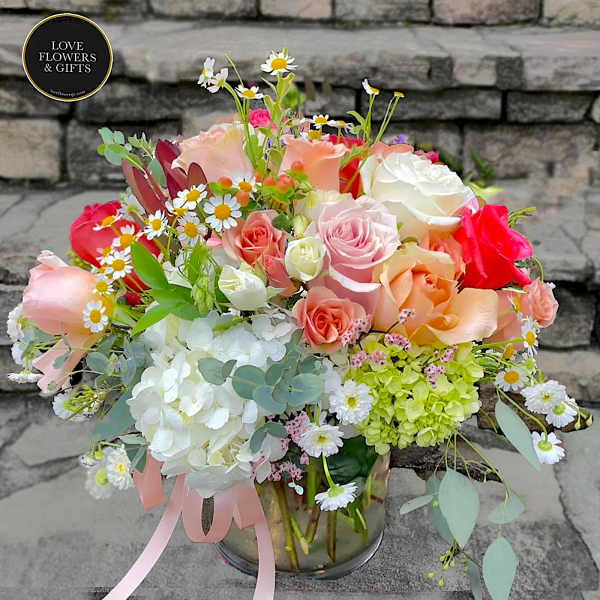 Low glass vase of mixed roses, hydrangeas, and daisies in soft pink, peach, white, and green tones