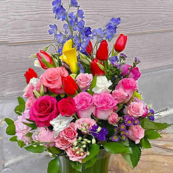 Mixed bouquet of pink roses, red tulips, yellow calla lilies and blue flowers in a clear glass vase.