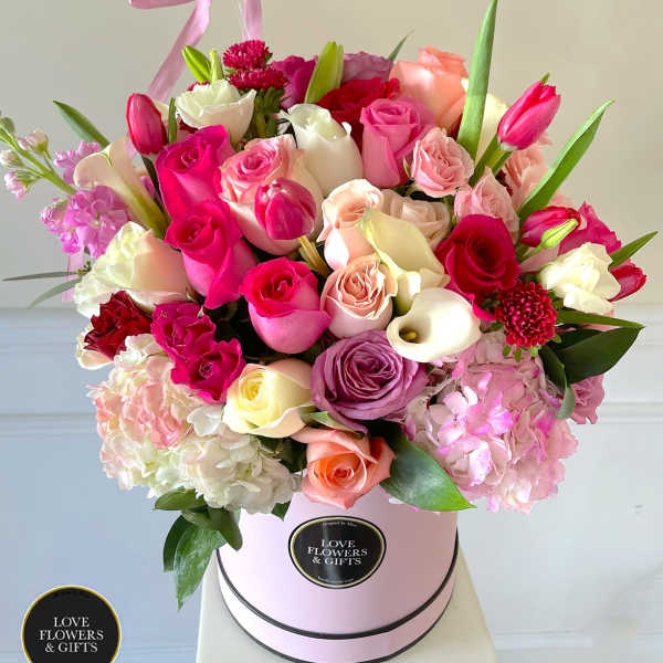 Round hatbox arrangement of mixed pink, white, and peach roses with tulips and calla lilies.