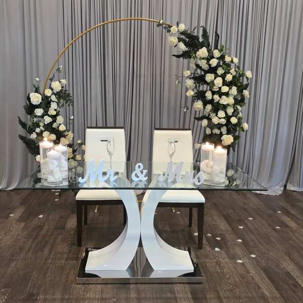 White rose arch over a glass table with candles and "Mr & Mrs" sign