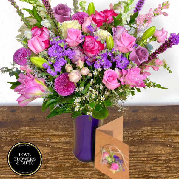 Tall arrangement of pink and purple roses, lilies, and mixed flowers in a purple vase with a kraft card.