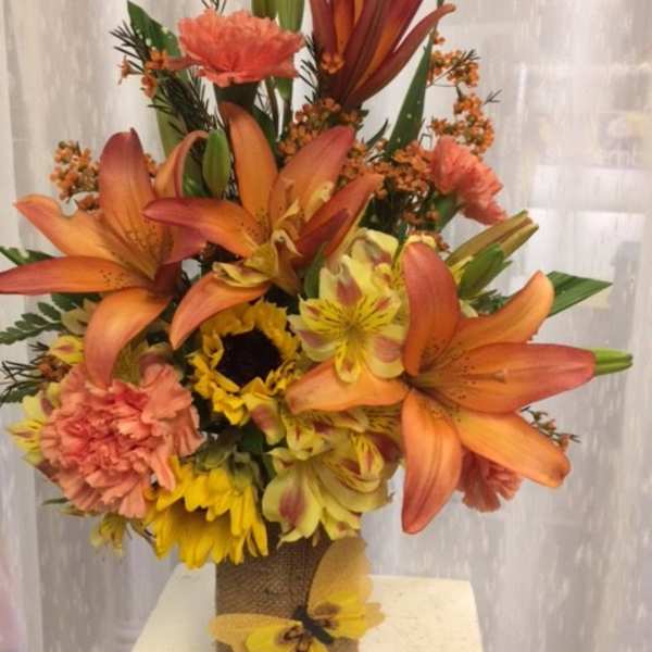 Tall bouquet of orange lilies, sunflowers, and carnations in a burlap-wrapped vase