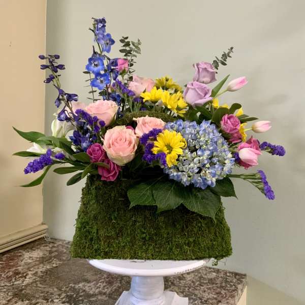 Mixed bouquet with roses, hydrangea, daisies, and purple blooms in a moss-covered container