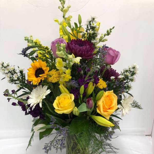 Tall mixed arrangement with yellow roses, sunflower, purple blooms and lilies in a clear glass vase