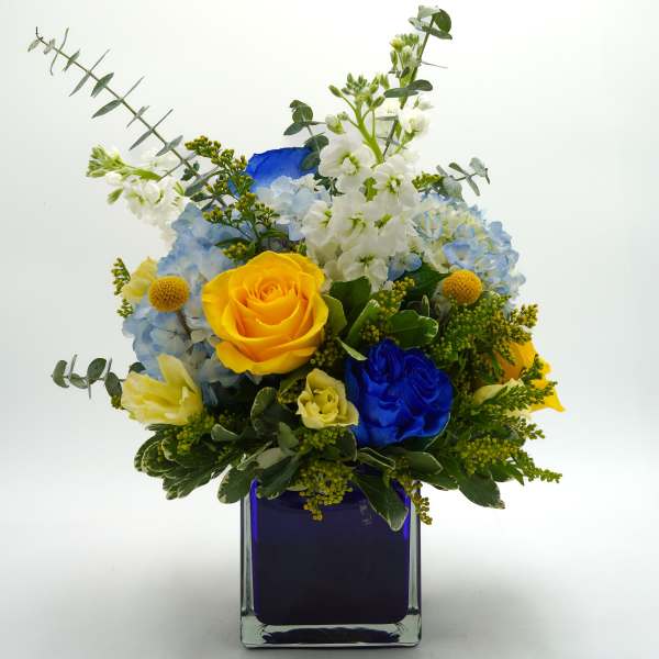 Bouquet of yellow and blue roses with white flowers in a square glass vase