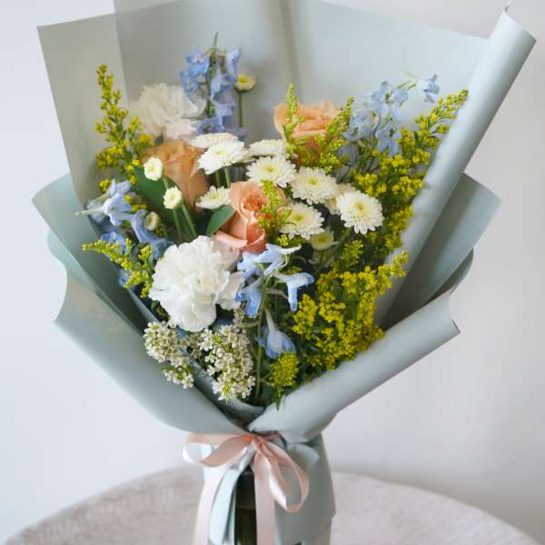Bouquet of peach roses, white daisies, and blue flowers wrapped in pale paper in a glass vase