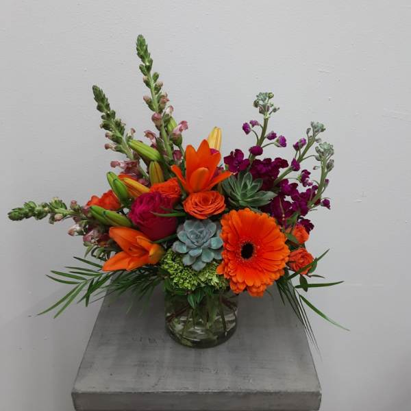 Bright mixed arrangement with orange gerbera, lilies, roses, and succulents in a clear glass vase