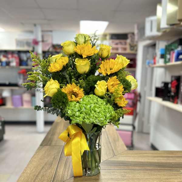 Yellow roses and sunflowers in a clear glass vase with a yellow ribbon