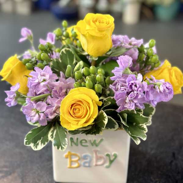 Yellow roses and lavender flowers in a white vase labeled New Baby