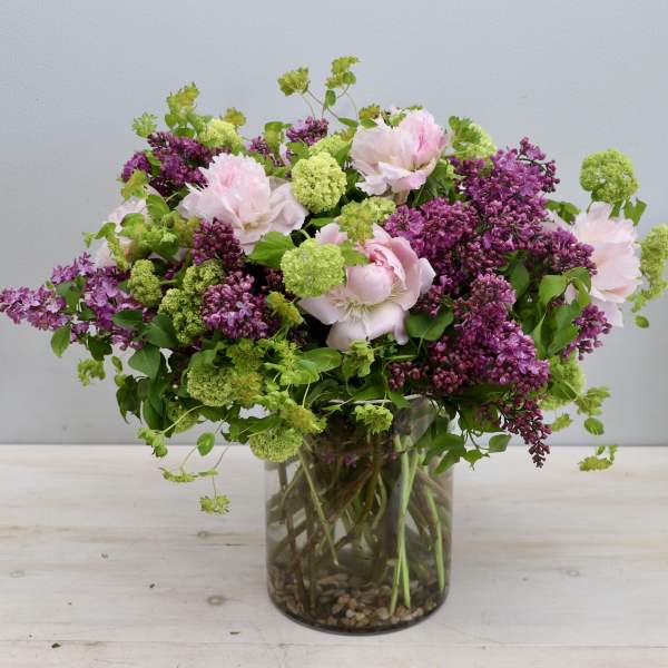 Loose garden-style bouquet of pink peonies, purple lilacs, and green blooms in a clear glass cylinder vase