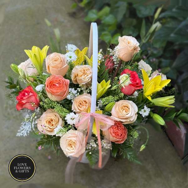 Basket arrangement of peach roses, red roses, and yellow lilies with a ribbon bow on the handle