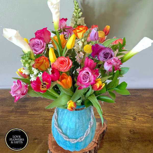 Colorful bouquet of roses, tulips, snapdragons, and calla lilies in a blue vase on a wood slab.
