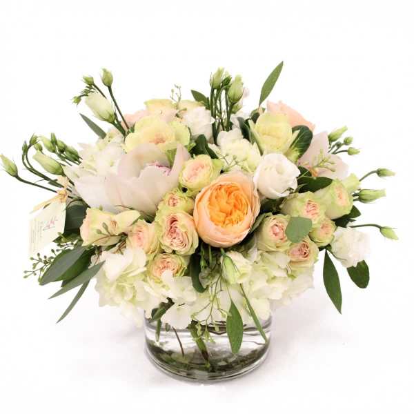 Low arrangement of white hydrangeas, peach roses, and cream blooms in a clear glass vase