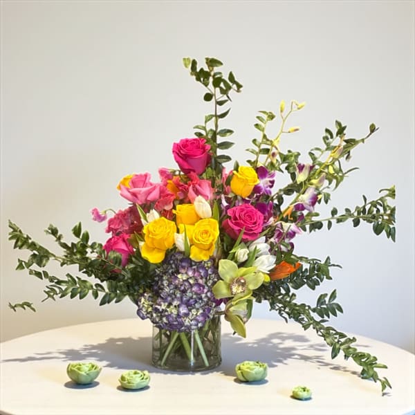 Bright arrangement of pink and yellow roses, orchids, tulips, and hydrangea in a glass vase with small succulents in cups.