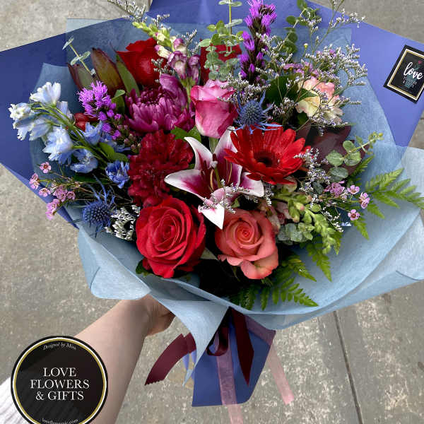 Hand-tied bouquet of red and pink roses, lilies, and mixed blooms wrapped in blue paper