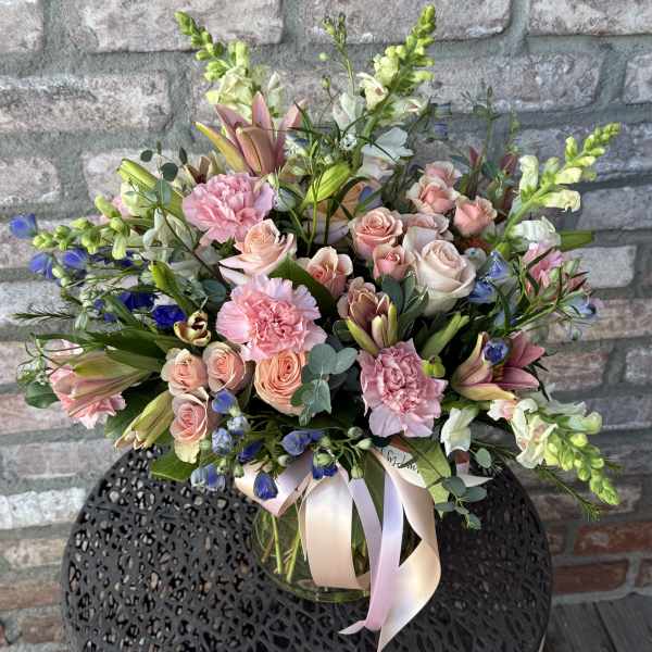 Mixed arrangement of pink roses, carnations, lilies, and blue flowers in a clear vase with a pink ribbon.