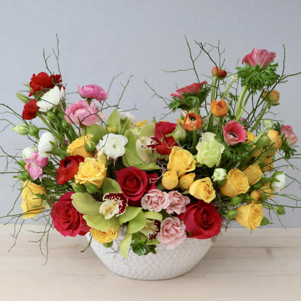 Low white bowl of mixed yellow, red, and pink roses with ranunculus and orchids in a loose, colorful arrangement